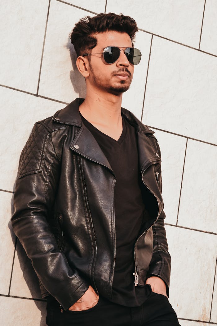 about-04 Fashionable young man in sunglasses and leather jacket posing against a tiled wall outdoors.