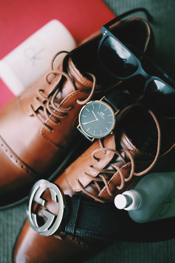 Brown leather shoes surrounded by classic men's accessories like a watch, belt, and sunglasses.